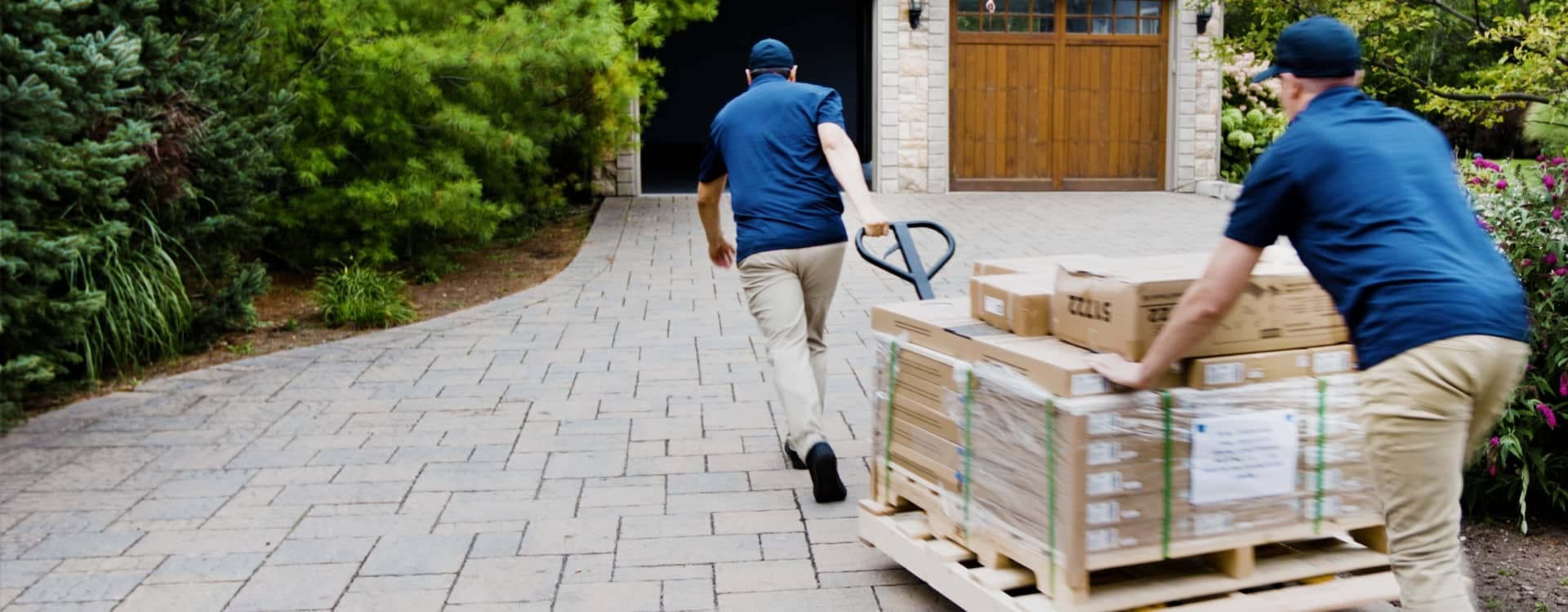 Two delivery workers wheeling boxes of flooring up a driveway to a home.