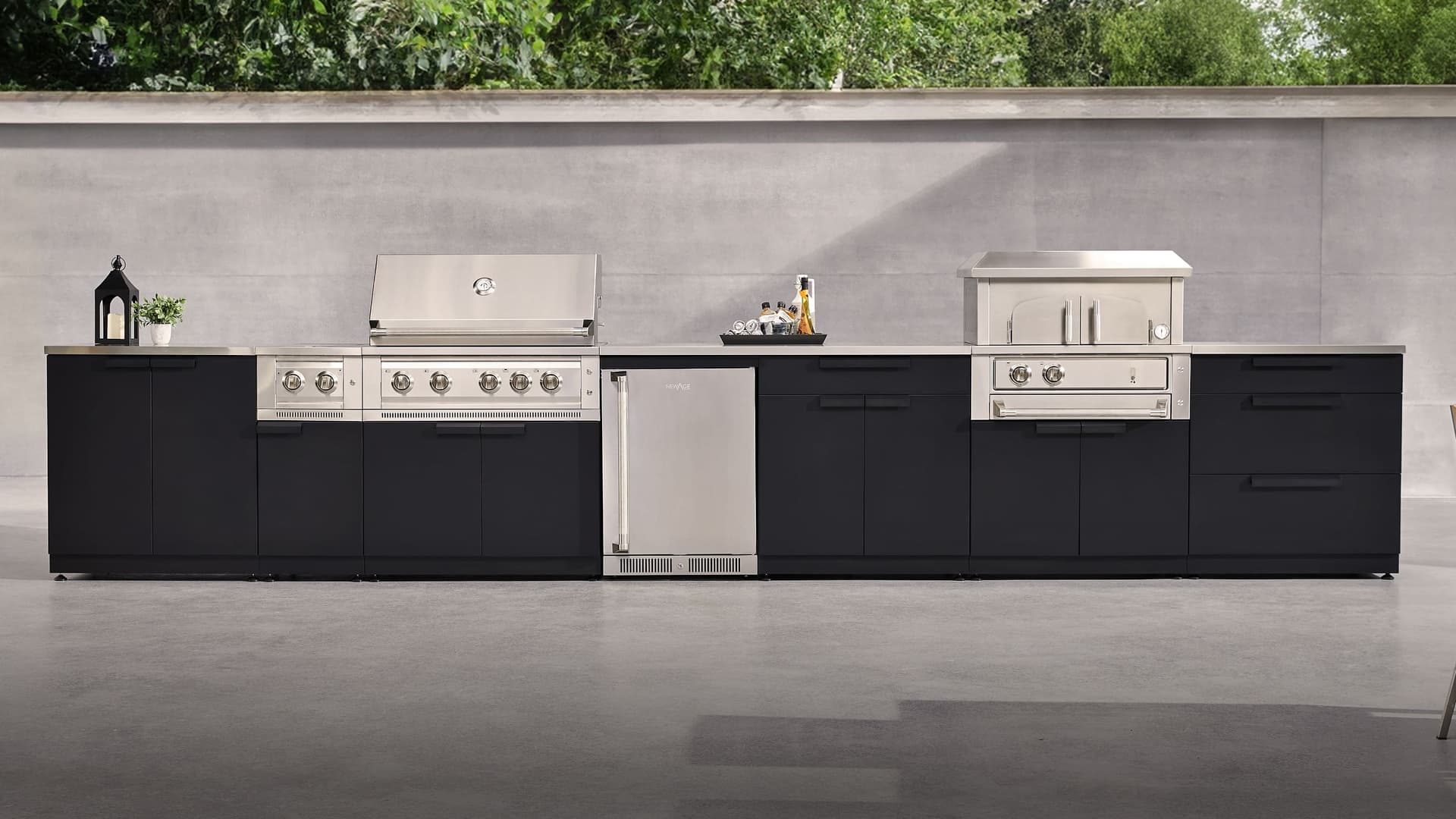 NewAge outdoor kitchen in matte black with a built-in gas grill, side burner, outdoor fridge and pizza oven against a concrete garden wall