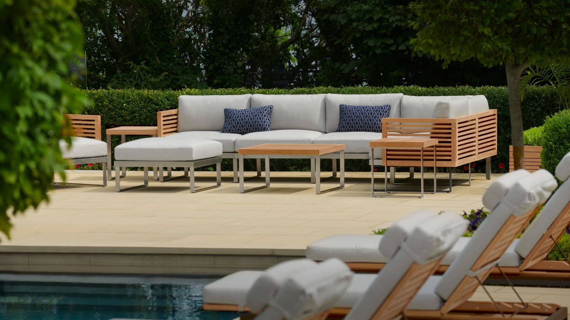 NewAge outdoor furniture set with a teak sectional, ottoman and coffee tables on a poolside patio, with loungers in the foreground
