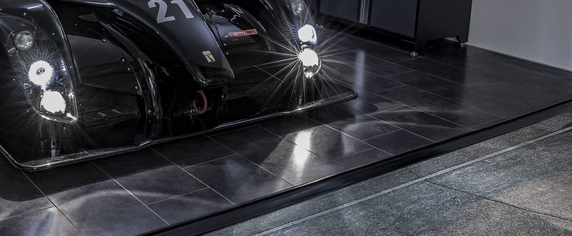 A black race car on glossy black vinyl tile flooring, aluminum strip joining a gray floor