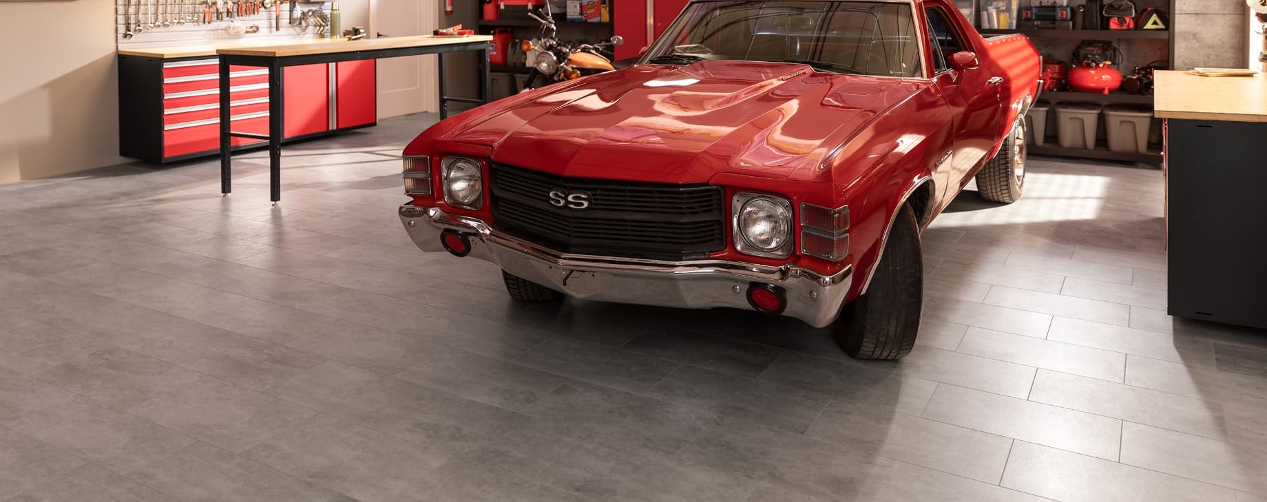 A red vintage muscle car on gray tile flooring, with red cabinets, workbench and motorcycle
