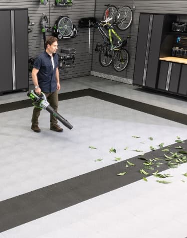 A man using a leaf blower on light gray floor tiles, with golf bags and wall-mounted bikes